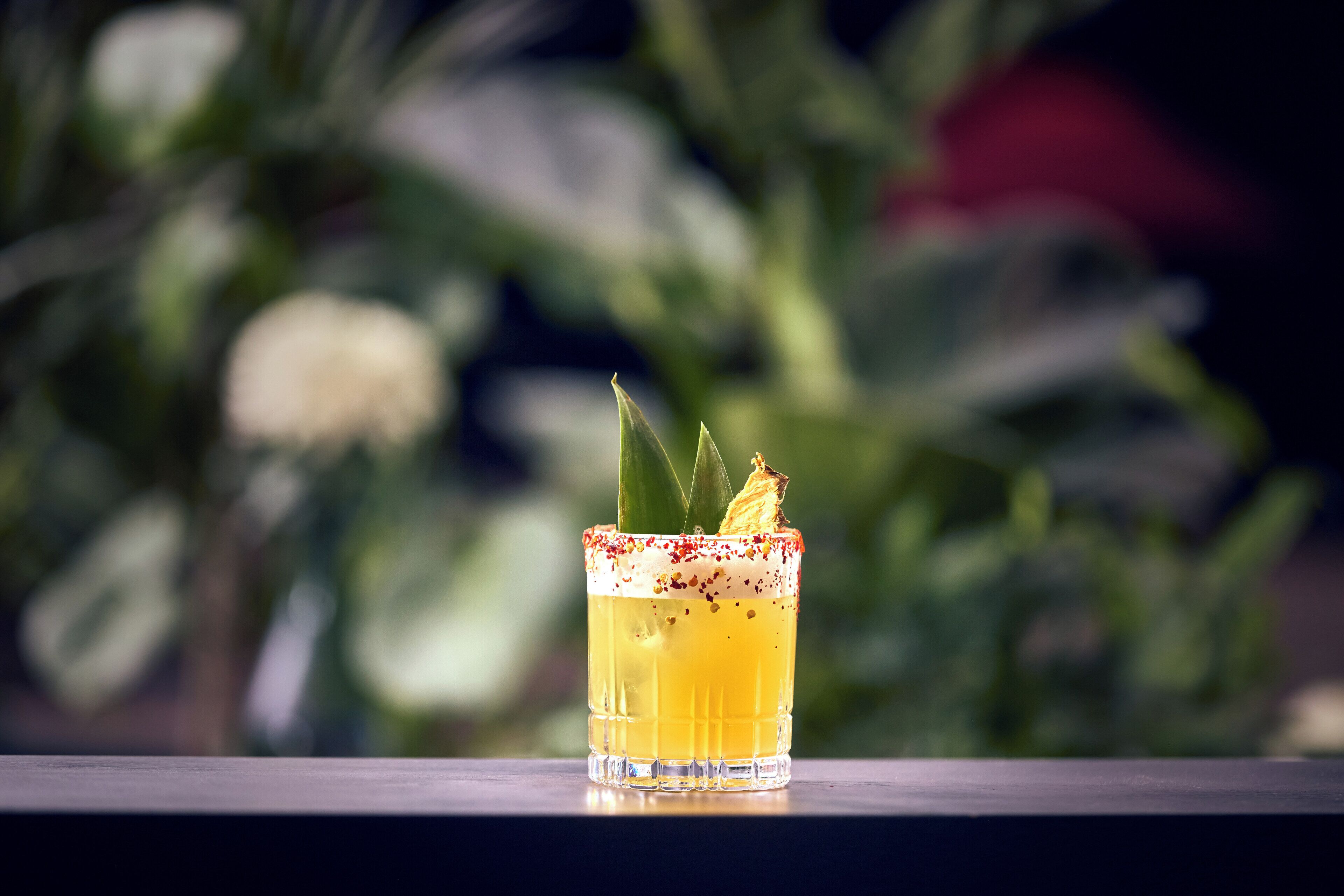 A cocktail with a garnish and a rim, placed on a table with a blurred background of greenery.