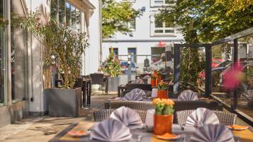 Ein Sitzbereich im Freien mit Tischen und Stühlen, Blumentöpfen und Blick auf die Straße.