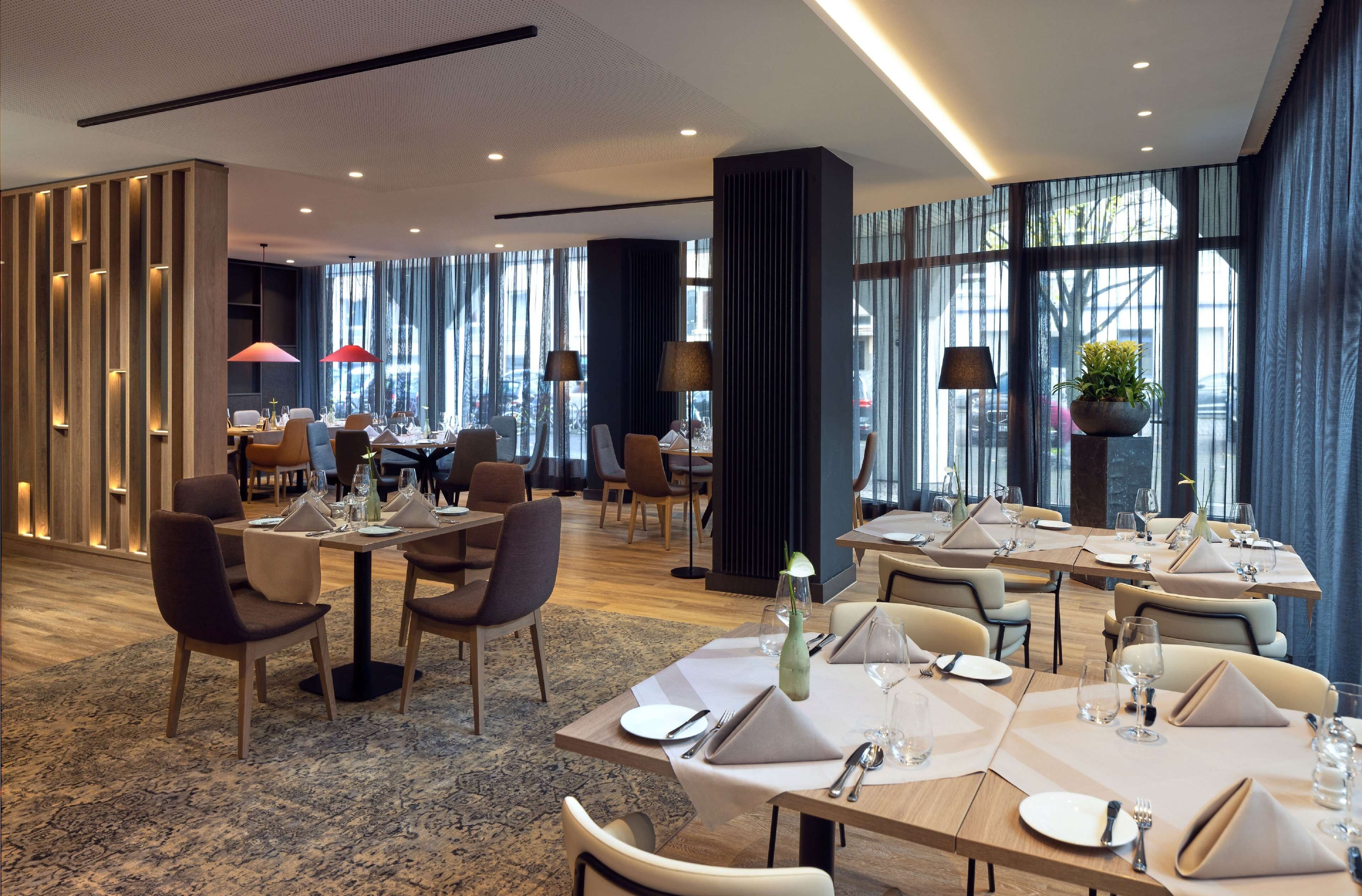 A modern hotel dining area with multiple tables set for guests, large windows offering views, and a contemporary interior design.