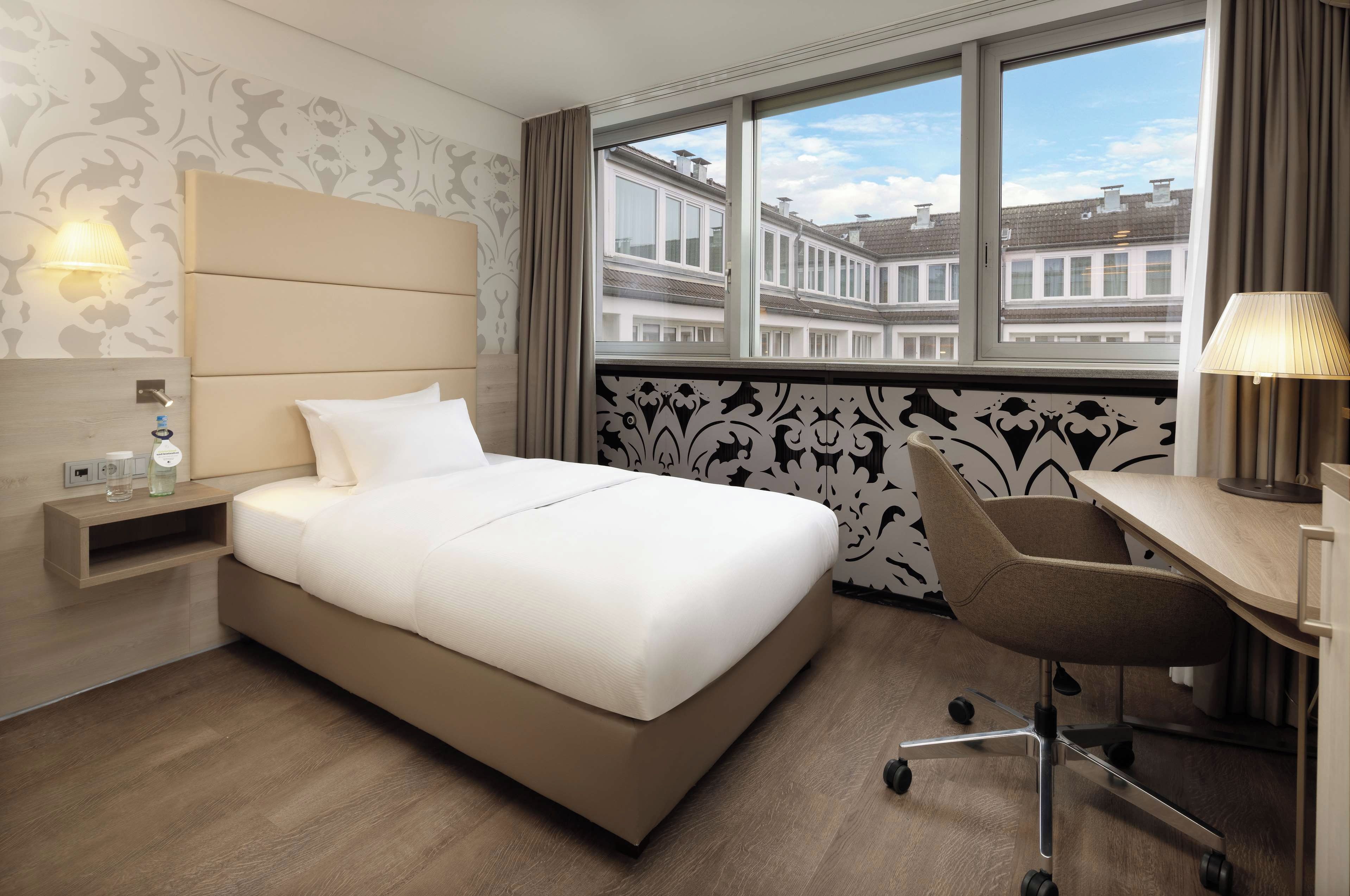 A hotel room with a bed, desk, chair, lamp, nightstand, and a view of buildings through the window.