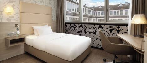 A hotel room with a bed, desk, chair, lamp, nightstand, and a view of buildings through the window.