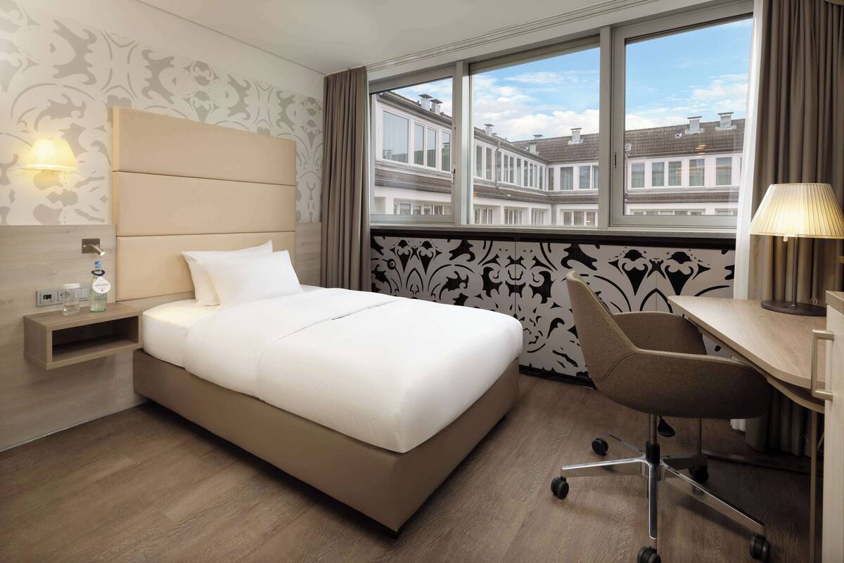 A hotel room with a bed, desk, chair, lamp, nightstand, and a view of buildings through the window.