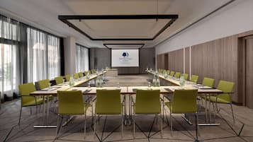 Un salón de conferencias con una larga mesa, sillas verdes y una pantalla proyectora que muestra