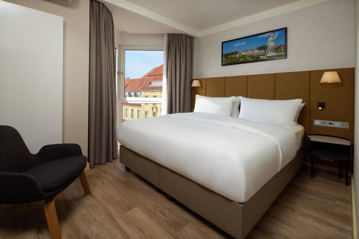 A hotel room with a large bed, a chair, a window with curtains, a painting, and a lamp.