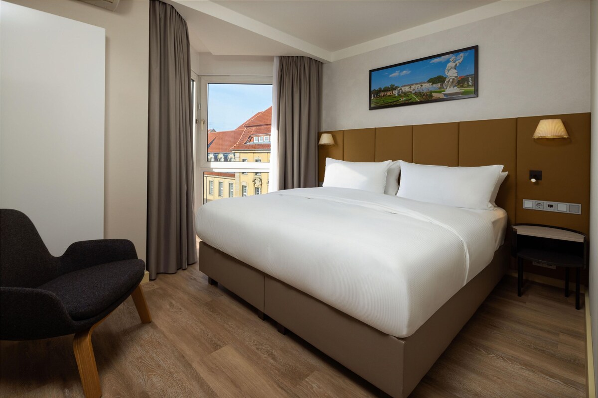 A hotel room with a large bed, a chair, a window with curtains, a painting, and a lamp.