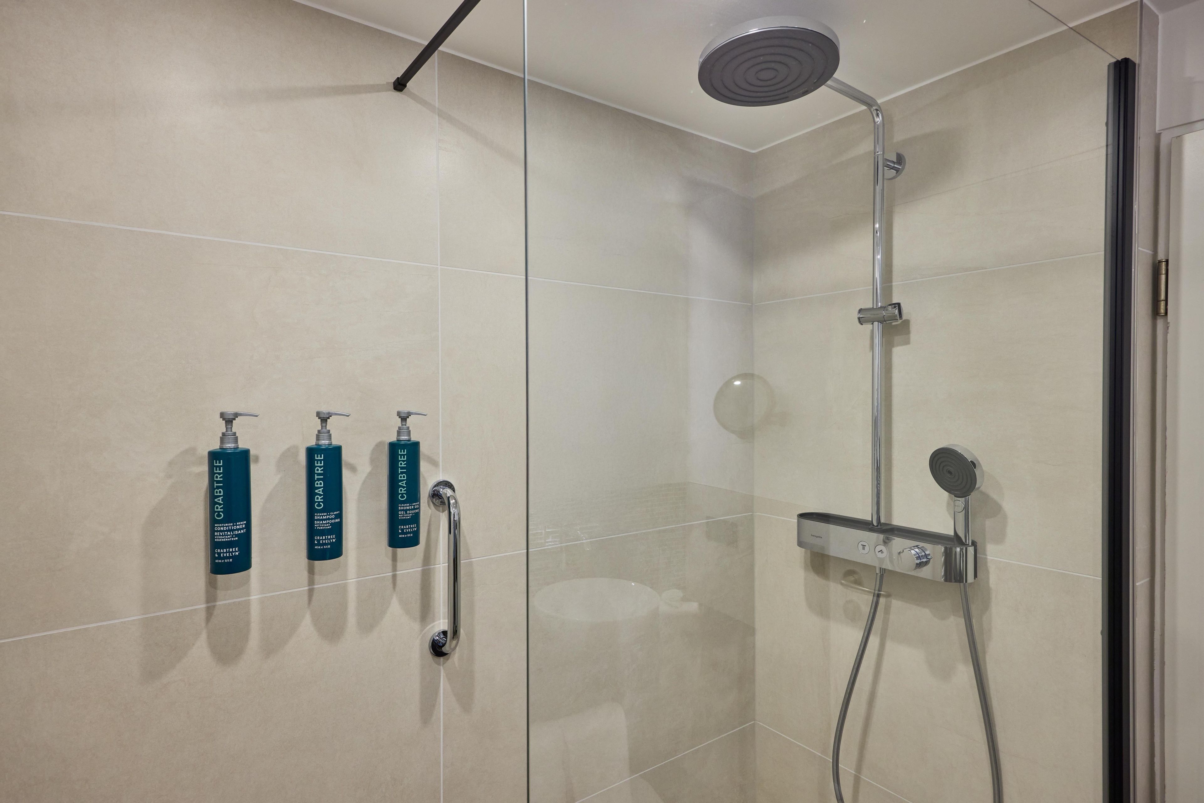 A modern bathroom with a glass shower enclosure, a handheld showerhead, and three bottles of Crabtree body wash.