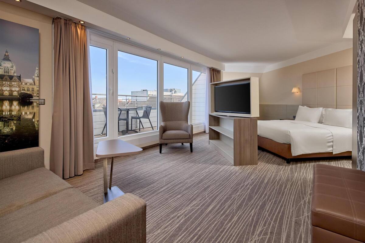A hotel room with a bed, a TV, a chair, a small table, and large windows offering a view of the city.