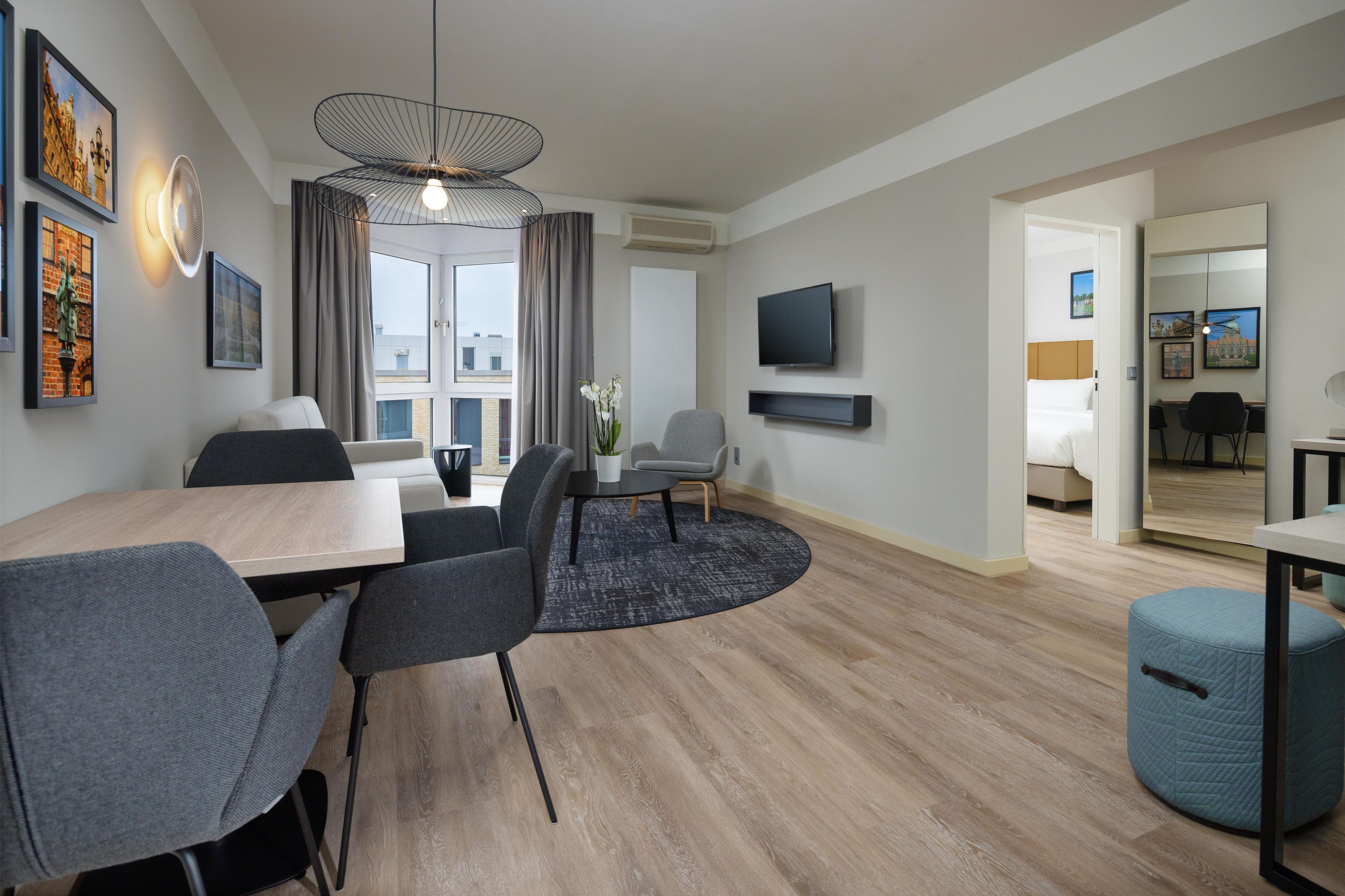 A modern hotel room with a dining area, a TV mounted on the wall, and a balcony with a view.