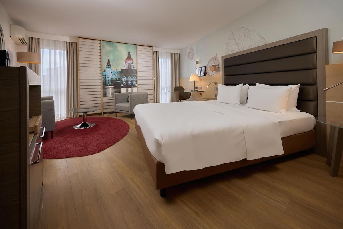 A hotel room with a large bed, a desk, a chair, a red rug, and a painting of a cityscape on the wall.