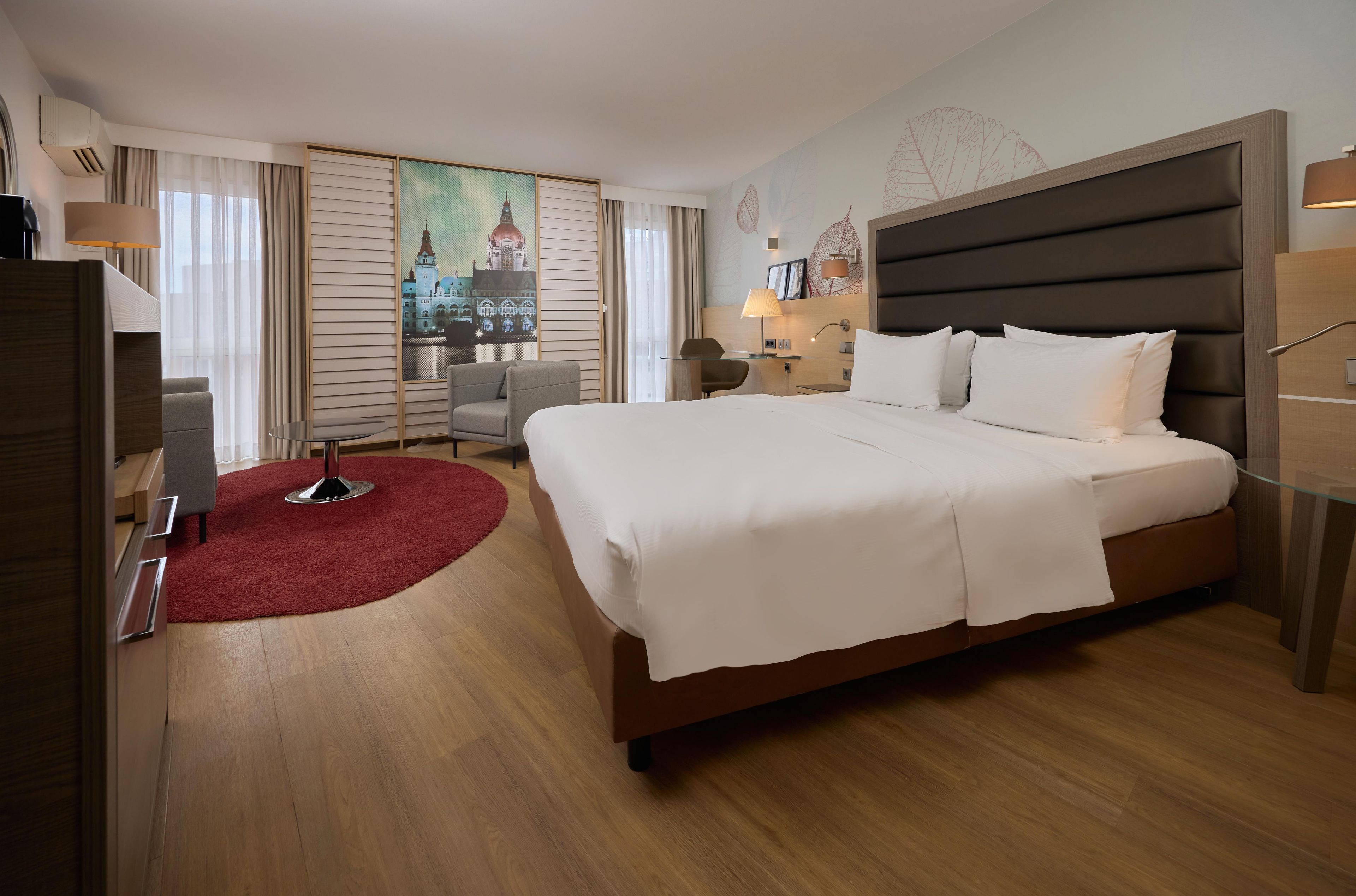 Ein Hotelzimmer mit einem großen Bett, einem Schreibtisch, einem Sessel, einem roten Teppich und einem Stadtbild an der Wand.