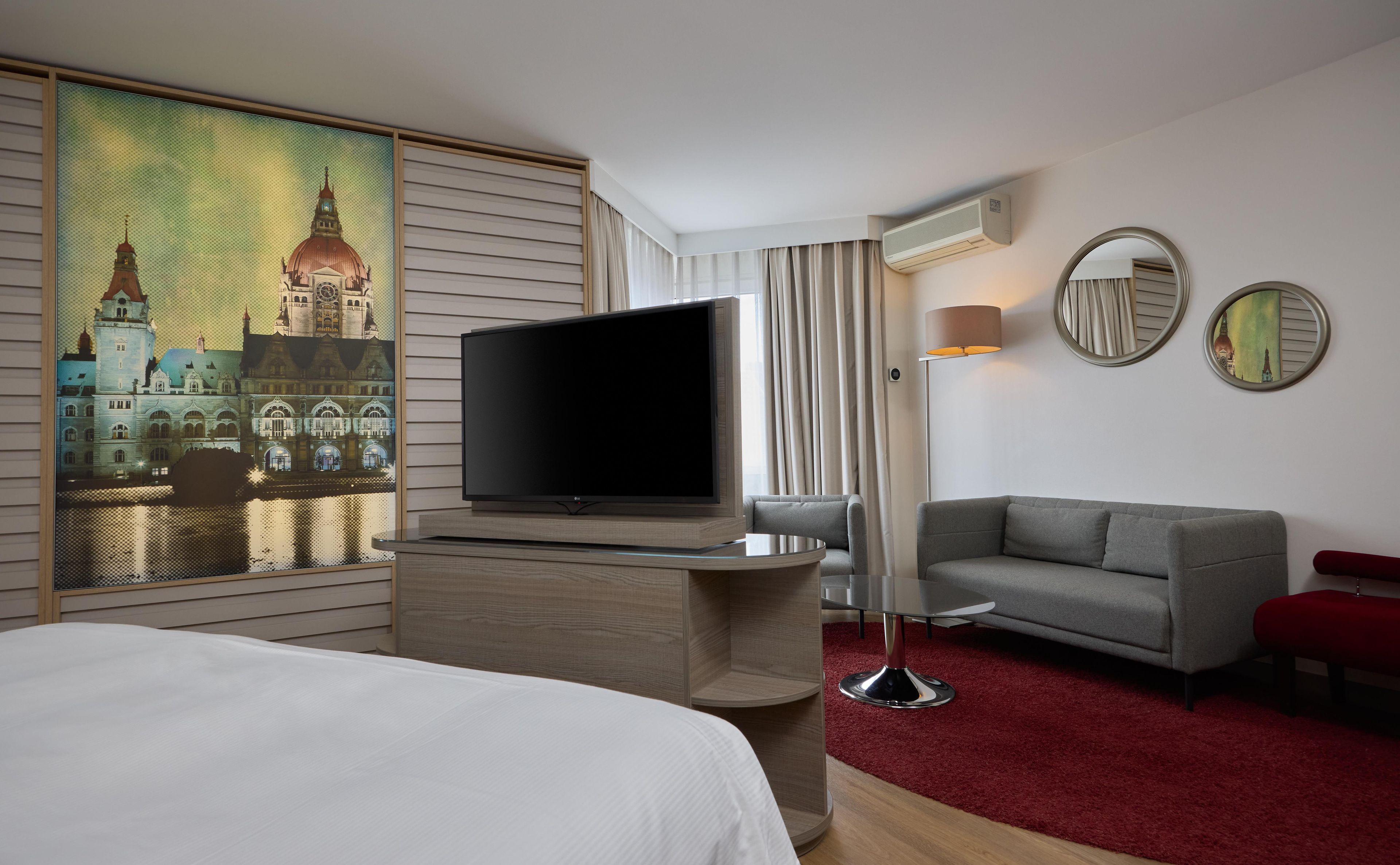 Une chambre d’hôtel dotée d’un grand téléviseur à écran plat, d’un canapé gris, d’un banc rouge et d’une moquette rouge.