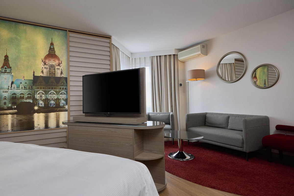 A hotel room with a large flat-screen TV, a grey sofa, a red bench, and a red carpet.