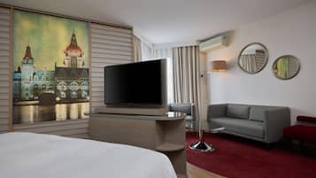 Une chambre d’hôtel dotée d’un grand téléviseur à écran plat, d’un canapé gris, d’un banc rouge et d’une moquette rouge.