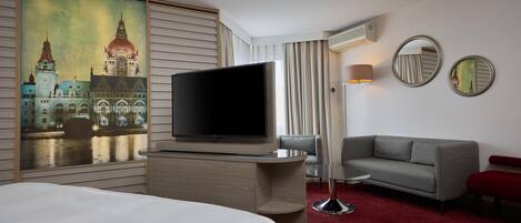 A hotel room with a large flat-screen TV, a grey sofa, a red bench, and a red carpet.