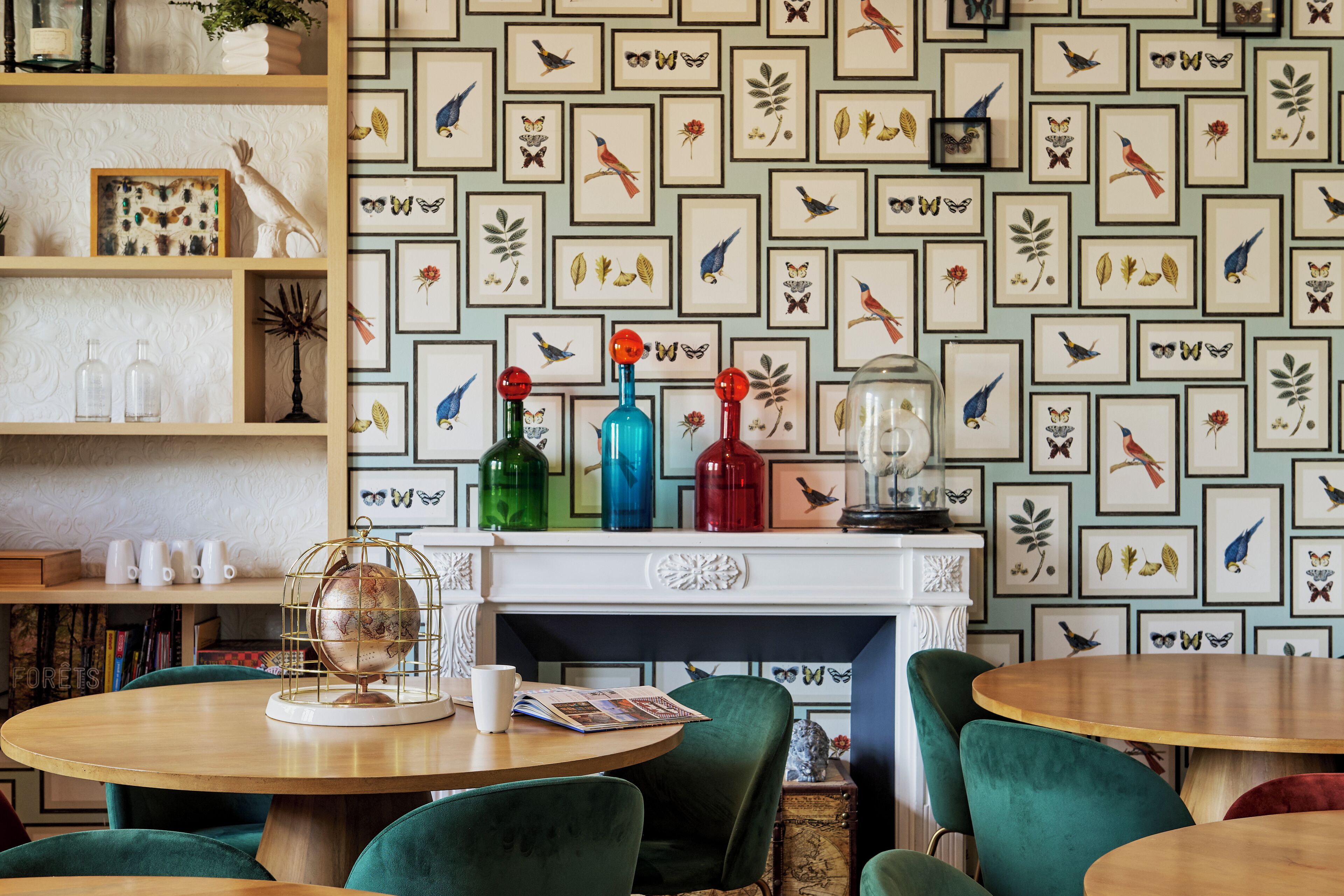 Une pièce chaleureuse avec une table ronde en bois, des fauteuils en velours vert et un papier peint à motifs représentant des oiseaux et des feuilles.
