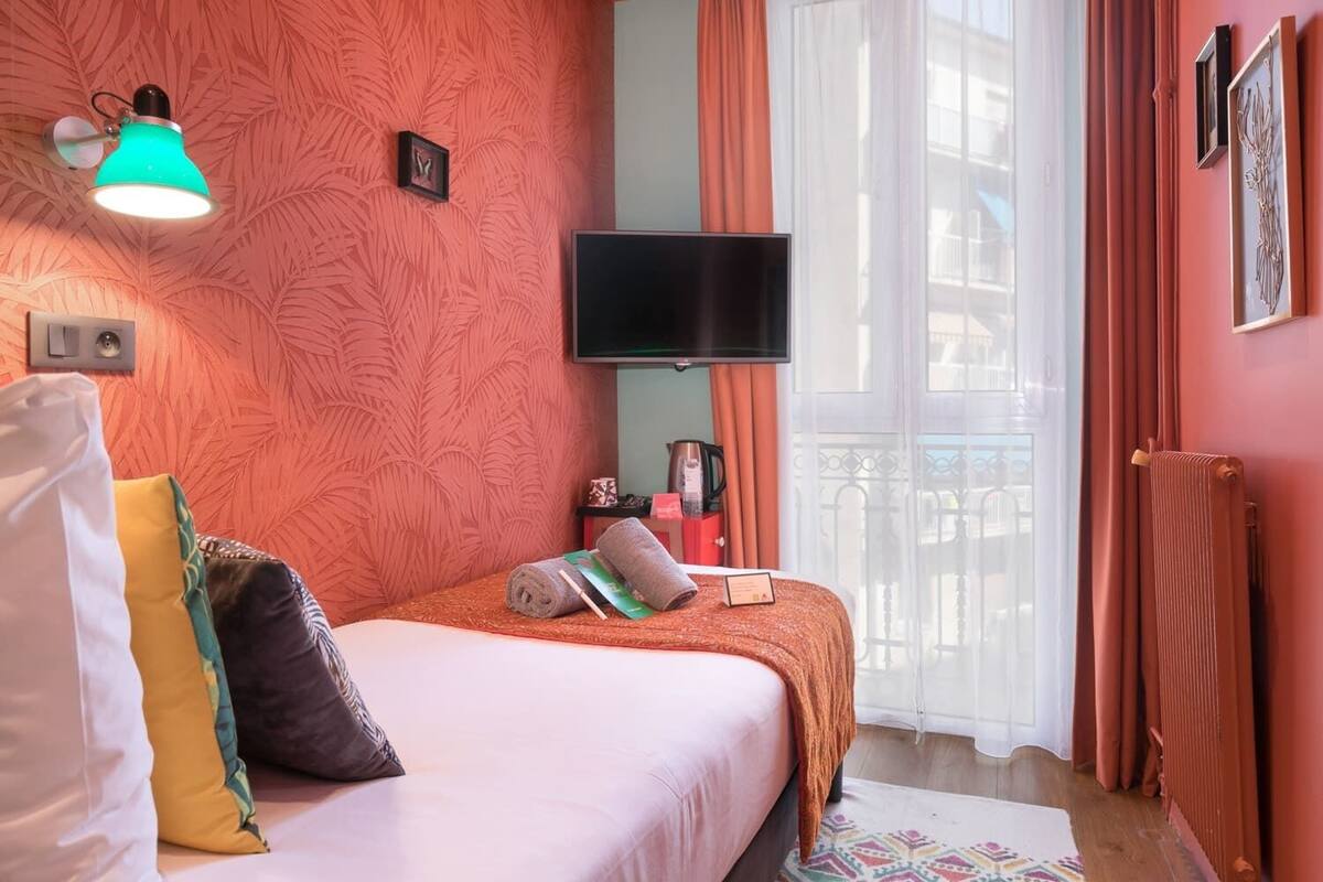 A hotel room with a bed, a television, a window with curtains, a radiator, and a wall-mounted light fixture.
