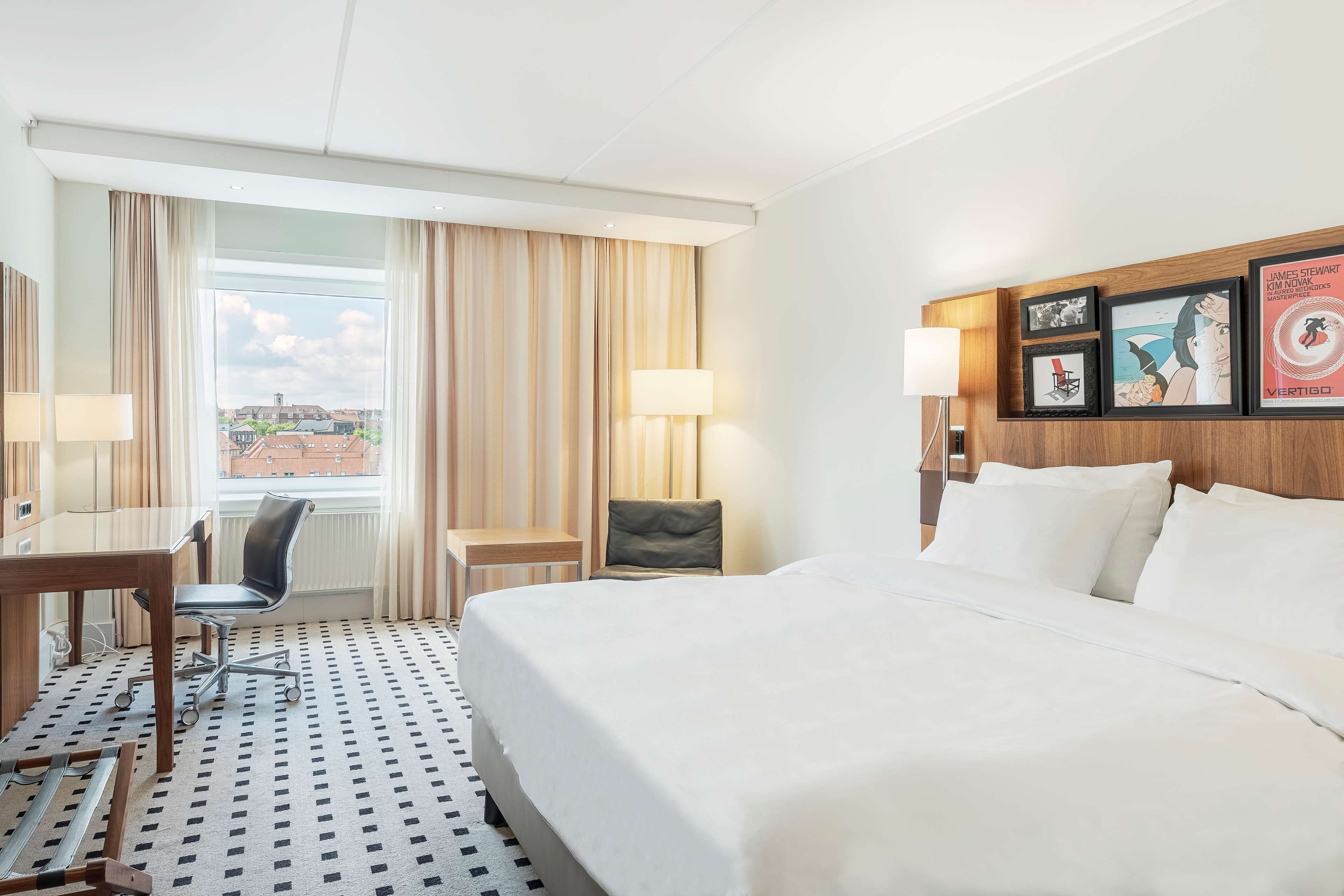 A hotel room with a bed, desk, chair, and a large window with curtains.
