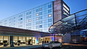 Un hotel moderno con fachada de vidrio, acceso cubierto y un coche estacionado frente a él.
