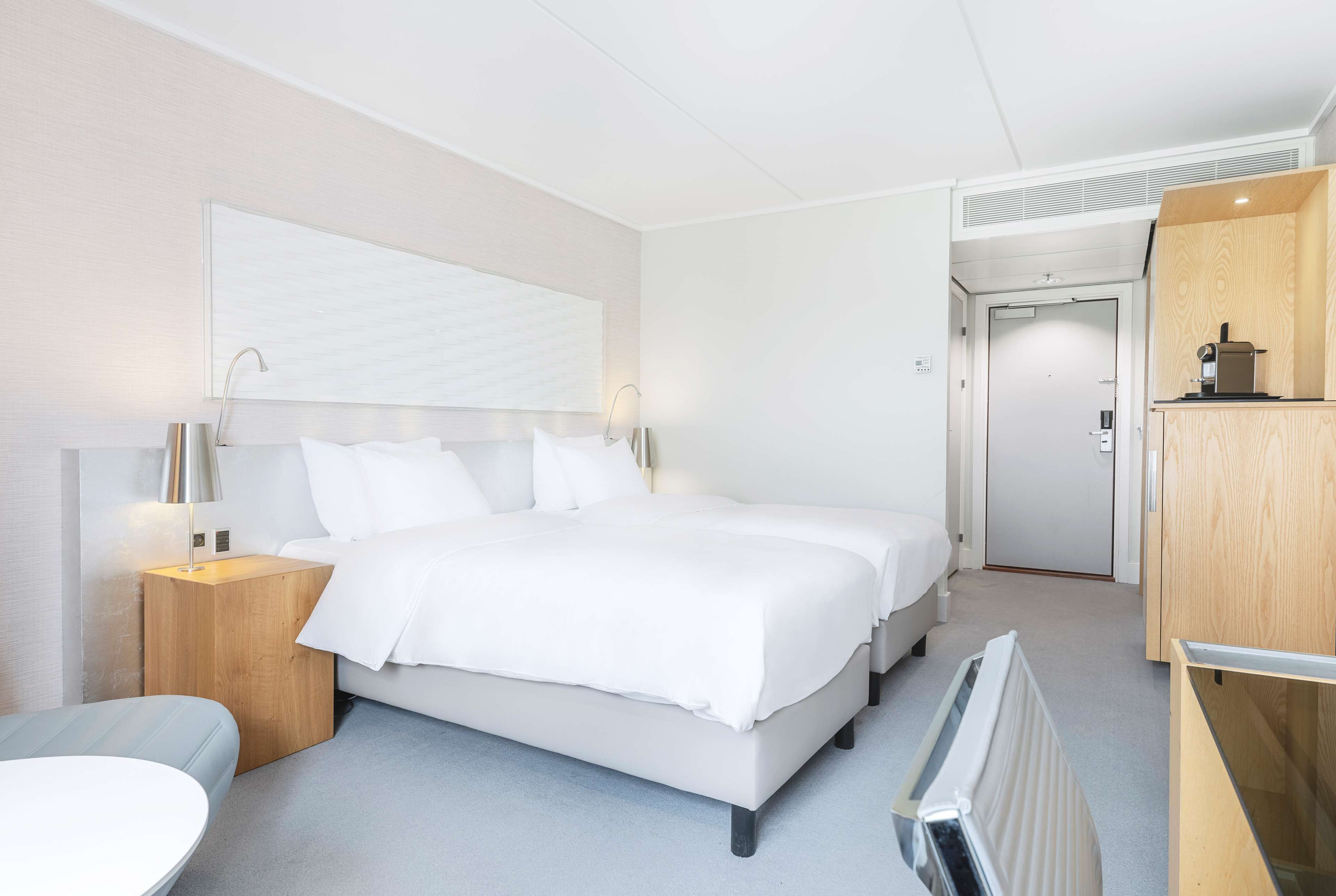A modern hotel room with a large bed, bedside tables, a wooden wardrobe, and a small kitchenette.