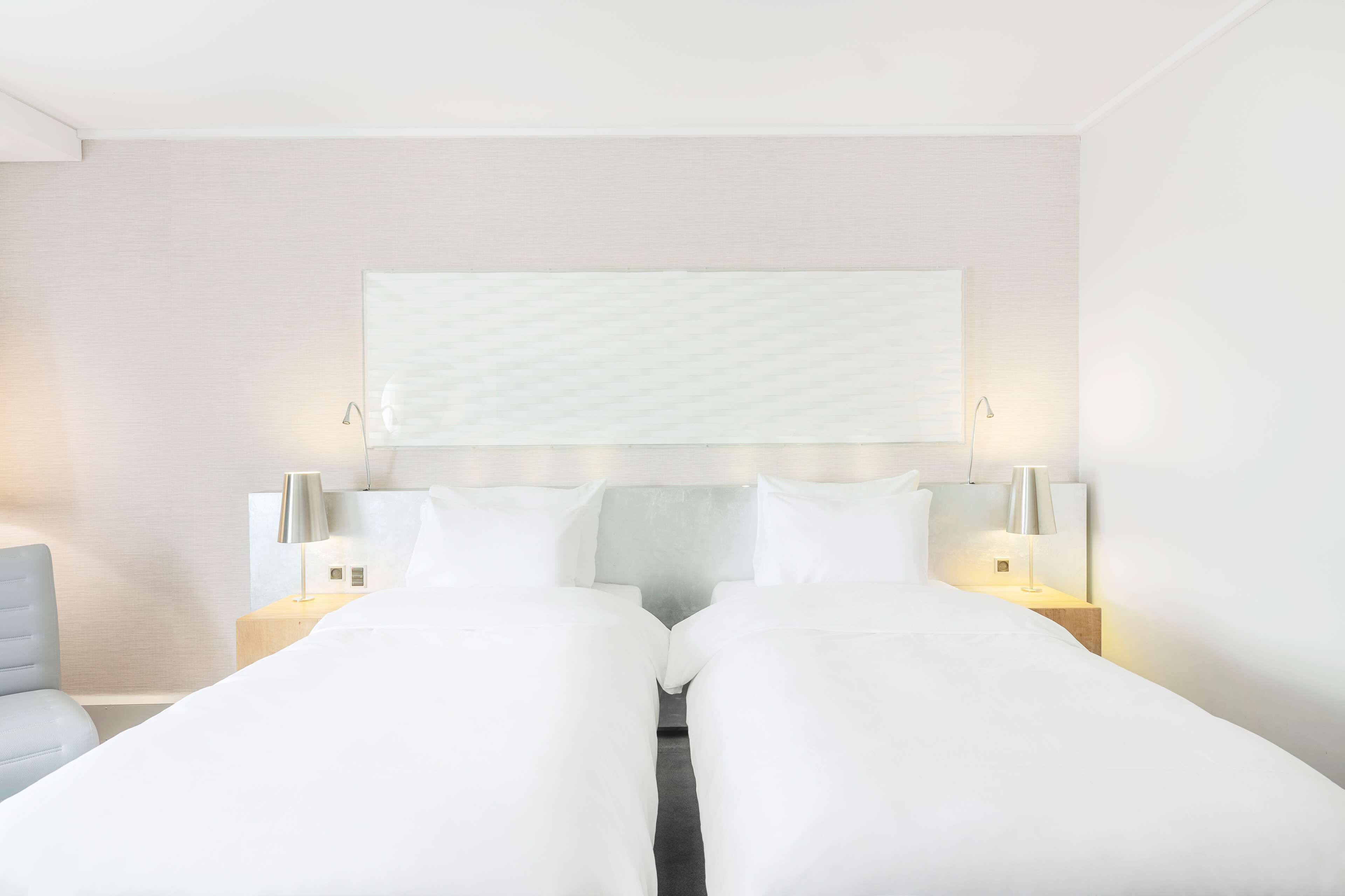 A hotel room with a large bed, two bedside tables with lamps, a headboard with a light panel, and a light-colored wall.