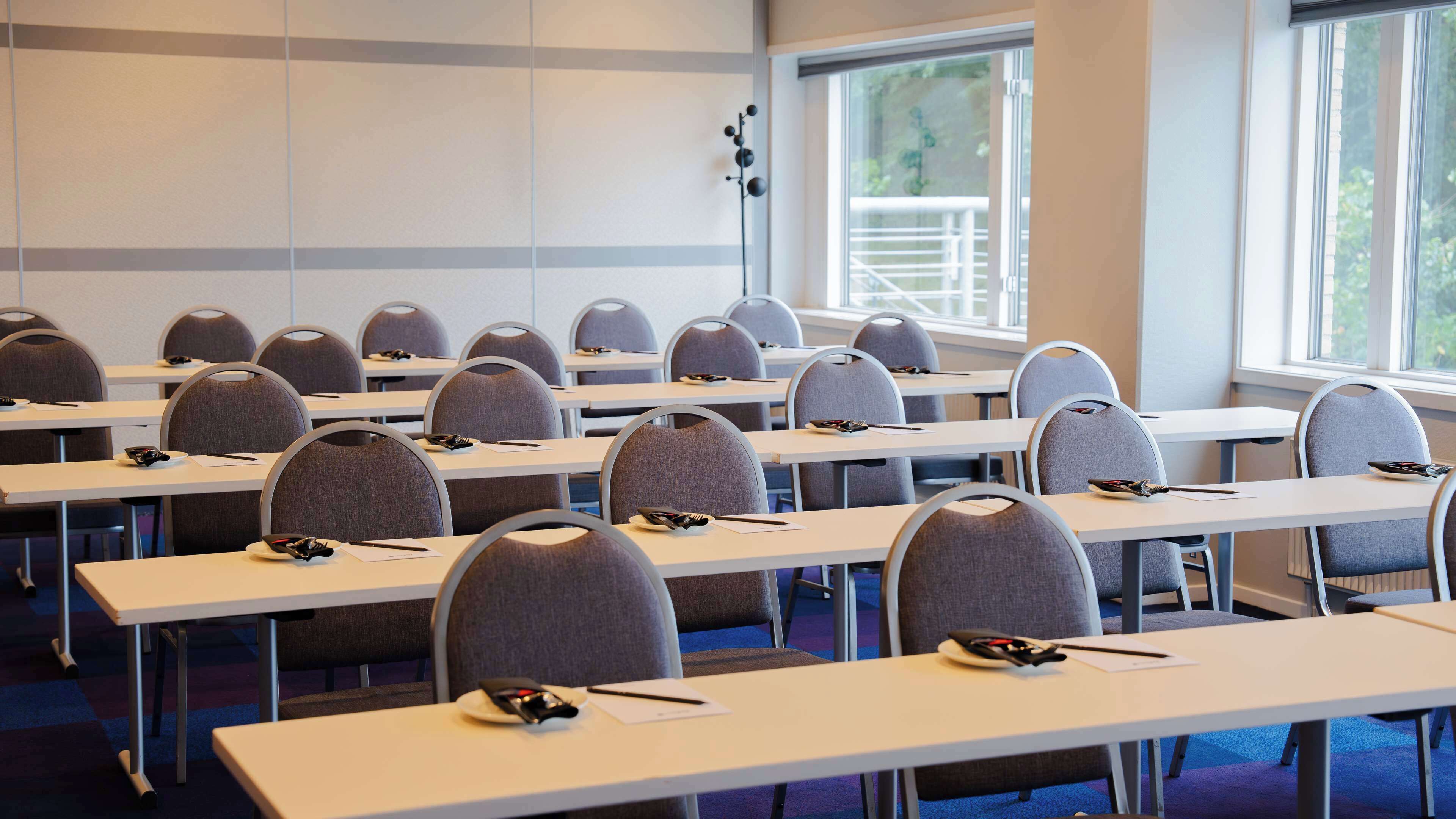 A conference room with rows of tables and chairs, large windows, and a view of greenery outside.