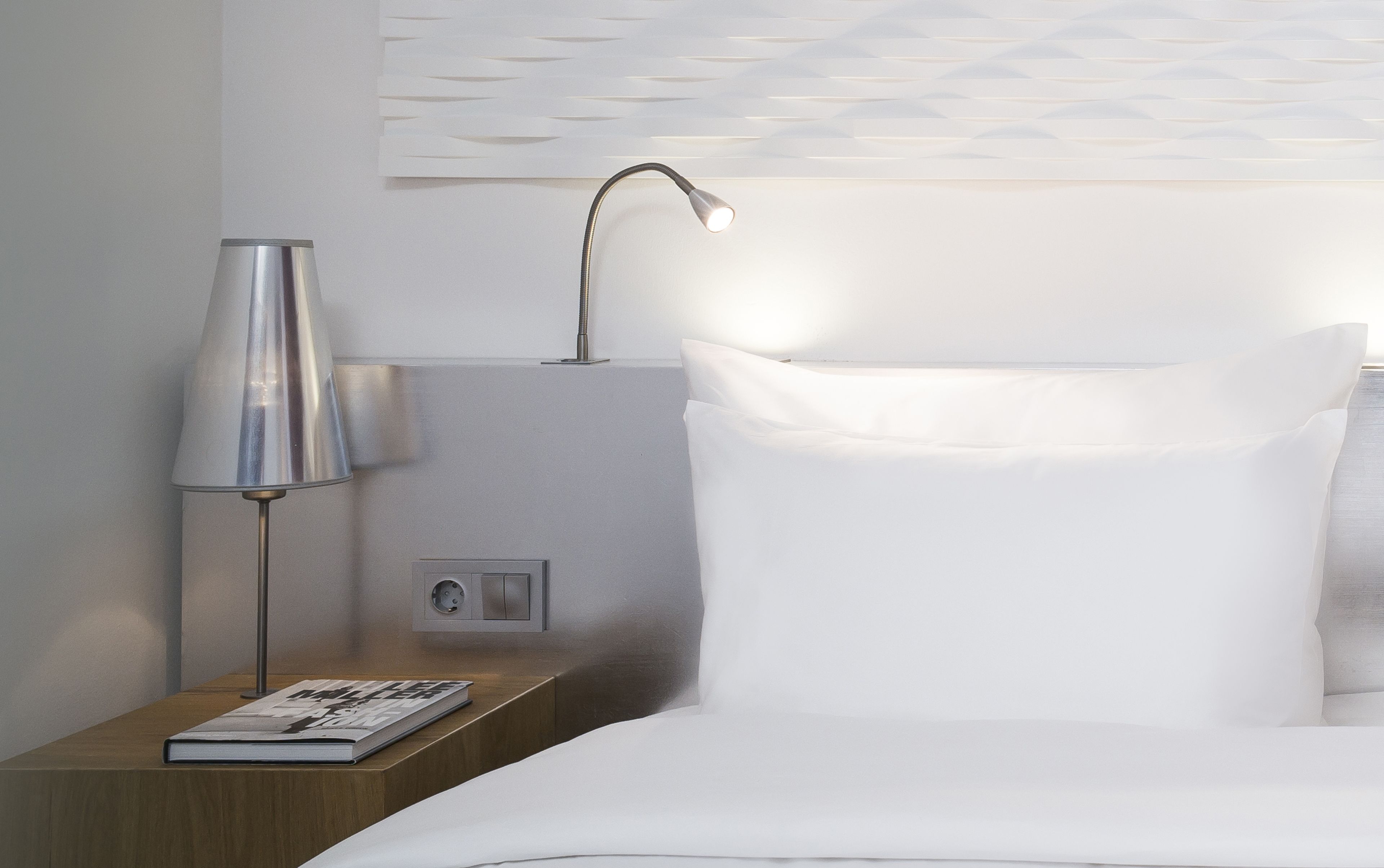 A neatly made bed with white linens, a bedside lamp, and a book on a wooden nightstand.