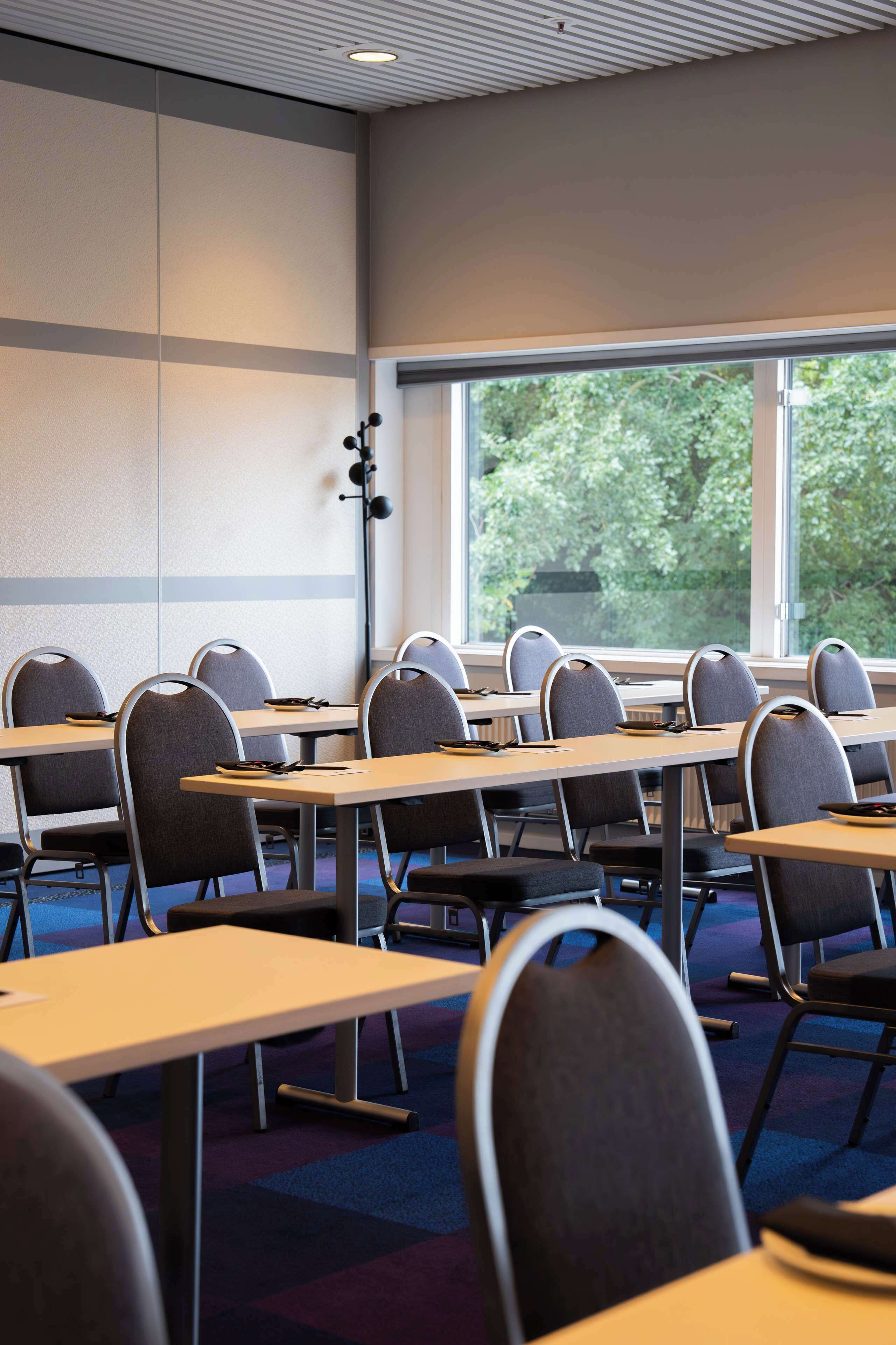 Un salón de conferencias con mesas y sillas, amplias ventanas con vistas a árboles e iluminación empotrada en el techo.