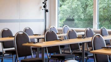 Un salón de conferencias con mesas y sillas, amplias ventanas con vistas a árboles e iluminación empotrada en el techo.