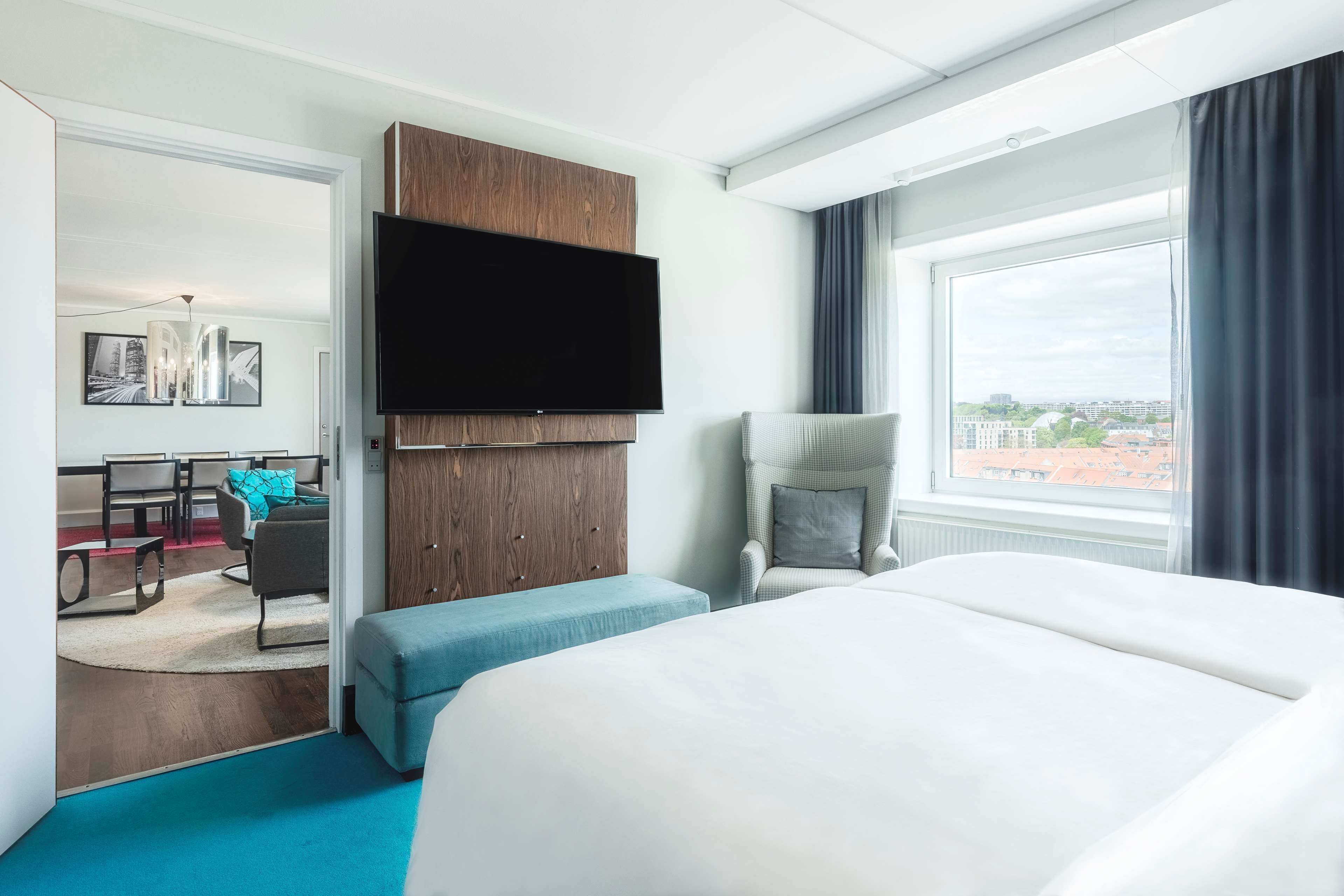 Habitación de hotel con una cama grande, televisión montada en un panel de madera, zona de estar con una silla y una mesa, y ventana con vista a la ciudad.