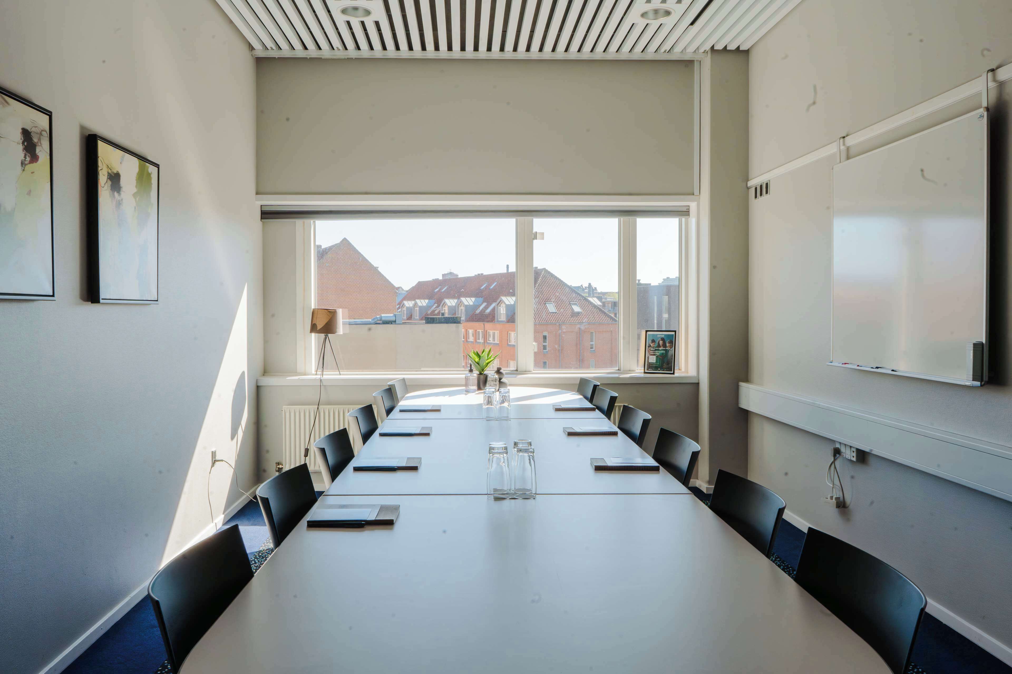 A conference room with a long table, chairs, a whiteboard, and a view of buildings through large windows.