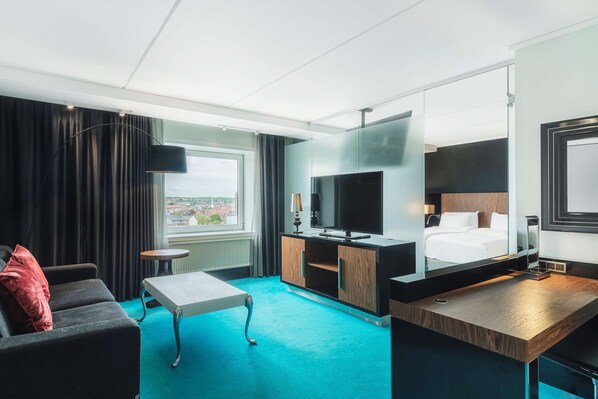 A modern hotel room with a large bed, a TV, a desk, and a view of the city through the window.