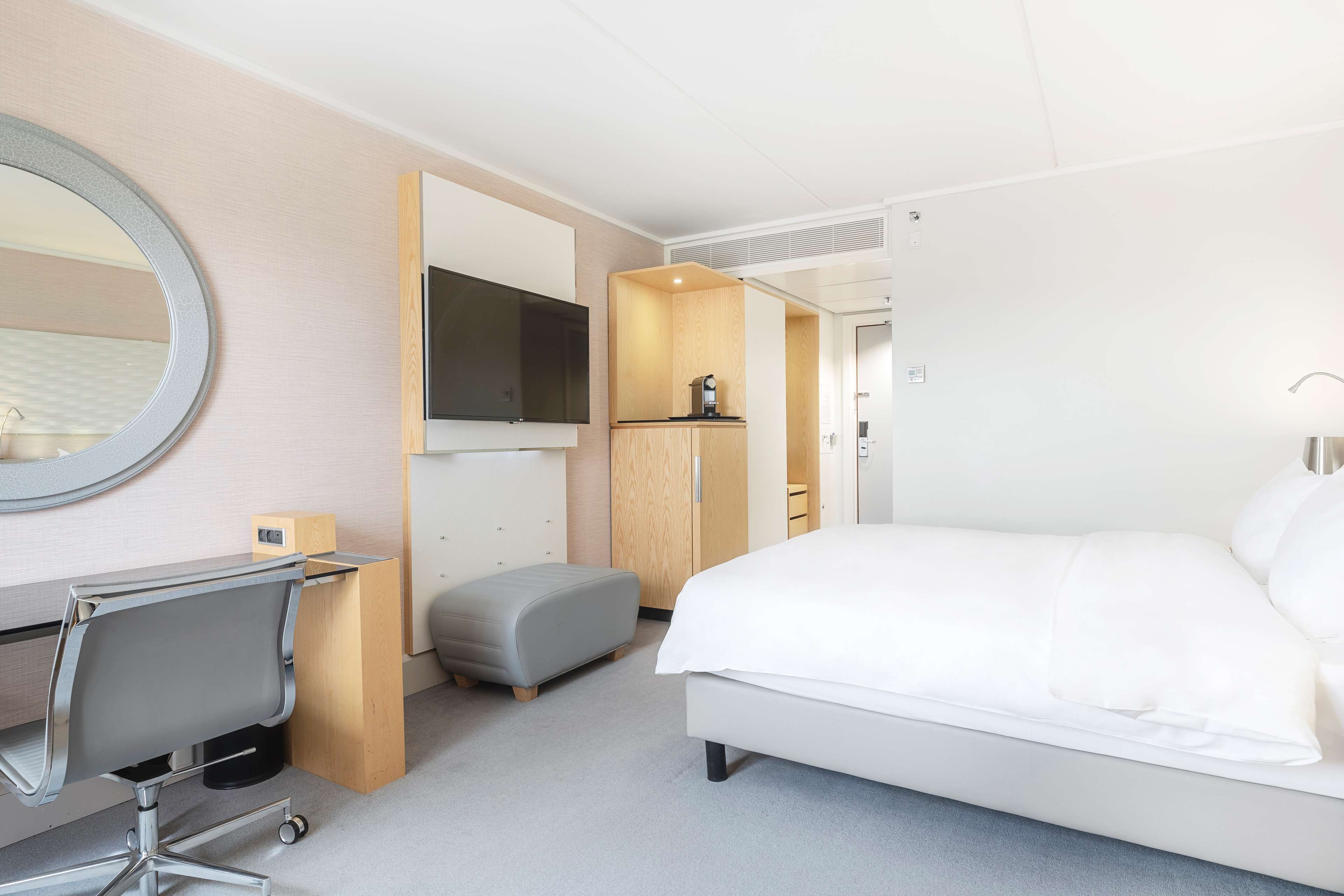 A hotel room with a bed, a desk with a chair, a small ottoman, a television, and a wooden cabinet.