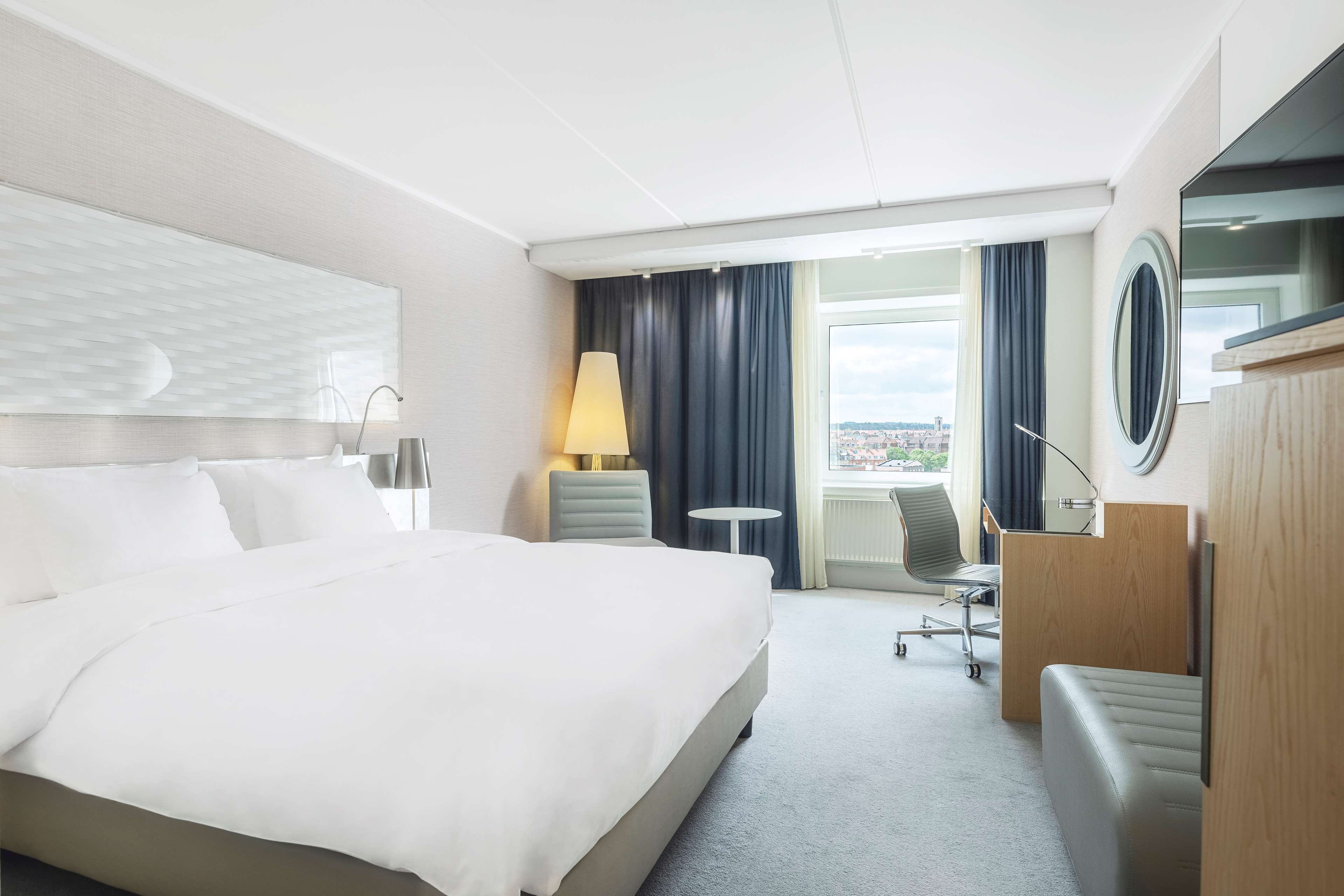 A hotel room with a large bed, a desk, a chair, and a view of the city.