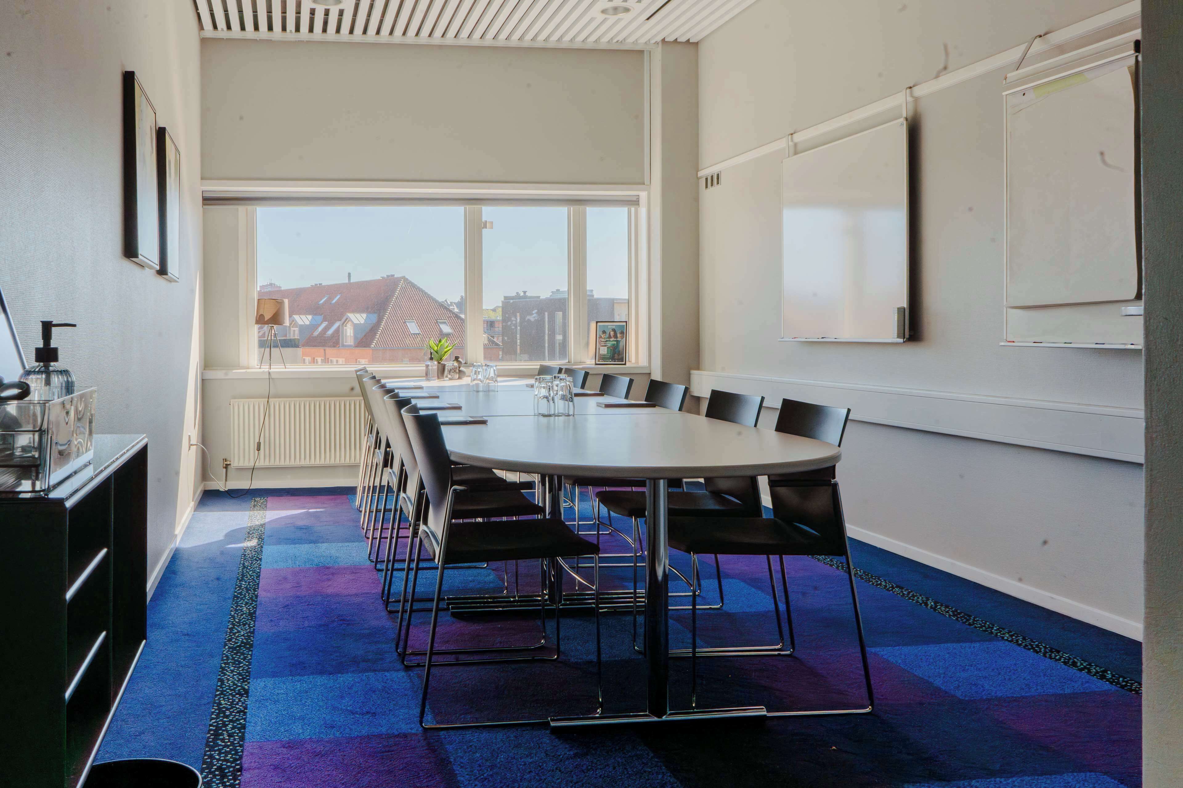Ein Konferenzraum mit großem Tisch, Stühlen, einer Whiteboard und Blick auf Gebäude durch ein großes Fenster.