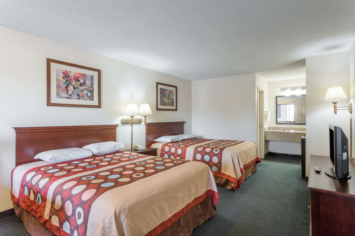 A hotel room with two beds, a desk, a TV, a lamp, and a bathroom visible in the background.