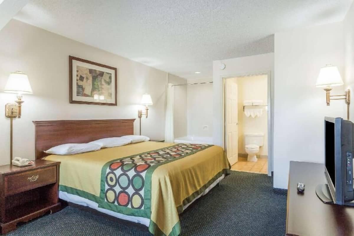 A hotel room with a bed, two bedside lamps, a TV, a painting on the wall, and a bathroom door.