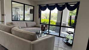 A modern living room with a beige sofa, a glass coffee table, and a large window with blue curtains. - Super 8 by Wyndham Slidell (Slidell)