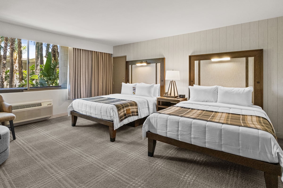 A hotel room with two beds, a window with curtains, a chair, and a bedside table with a lamp.