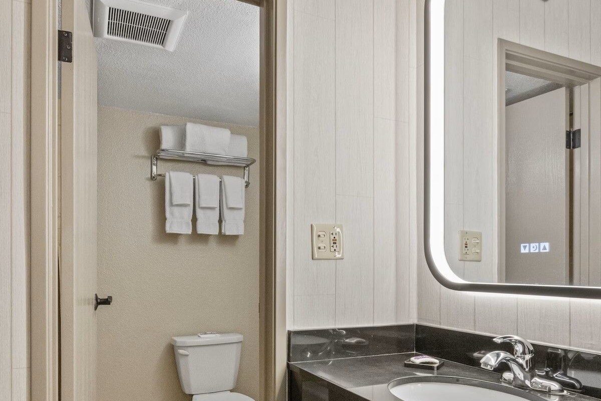 Un baño con inodoro, lavamanos y espejo. Hay un perchero sobre el inodoro y un cubo de basura al lado del lavamanos.