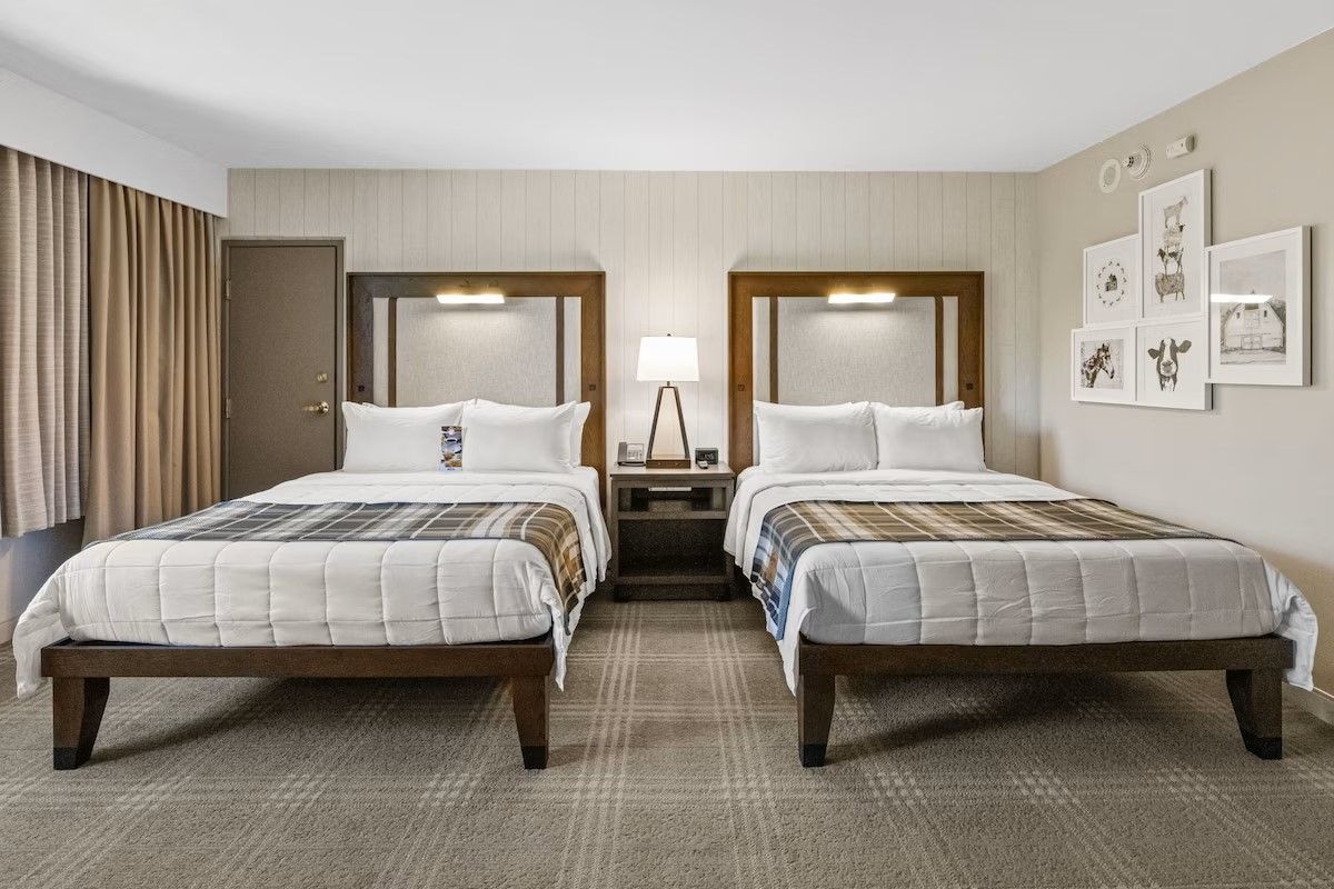 A hotel room with two beds, a nightstand, a lamp, and framed pictures on the wall.