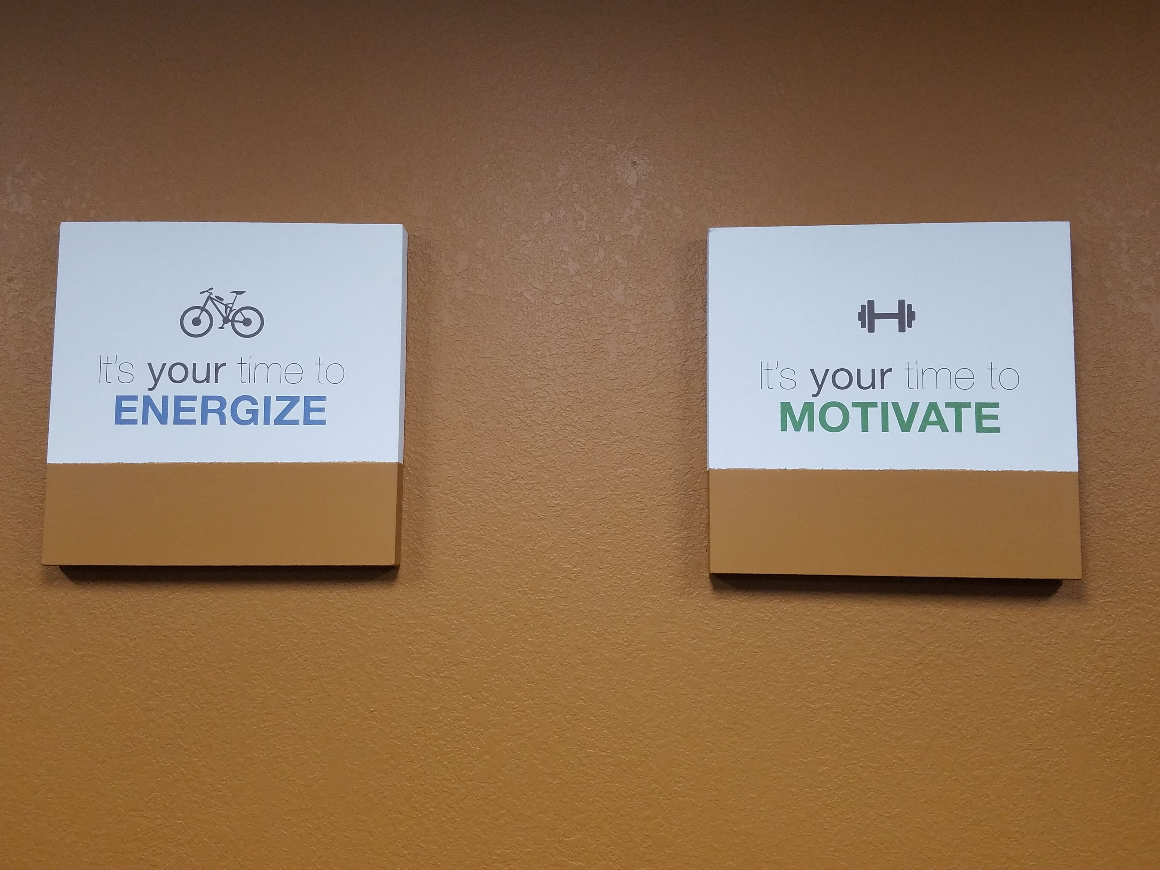 Two motivational signs on a wall, one encouraging to 'ENERGIZE' with a bicycle icon, and the other to 'MOTIVATE' with a dumbbell icon.