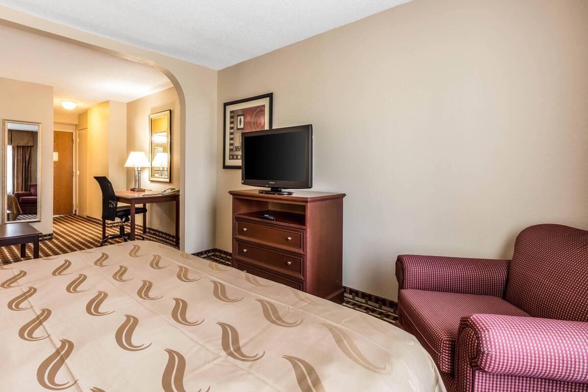 Quarto de hotel com uma cama, televisão, escrivaninha com cadeira e uma poltrona vermelha.