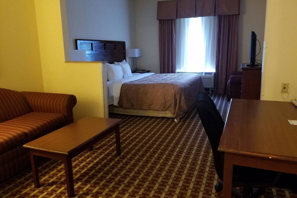 Quarto de hotel com cama, sofá, escrivaninha, televisão e janela com cortinas.
