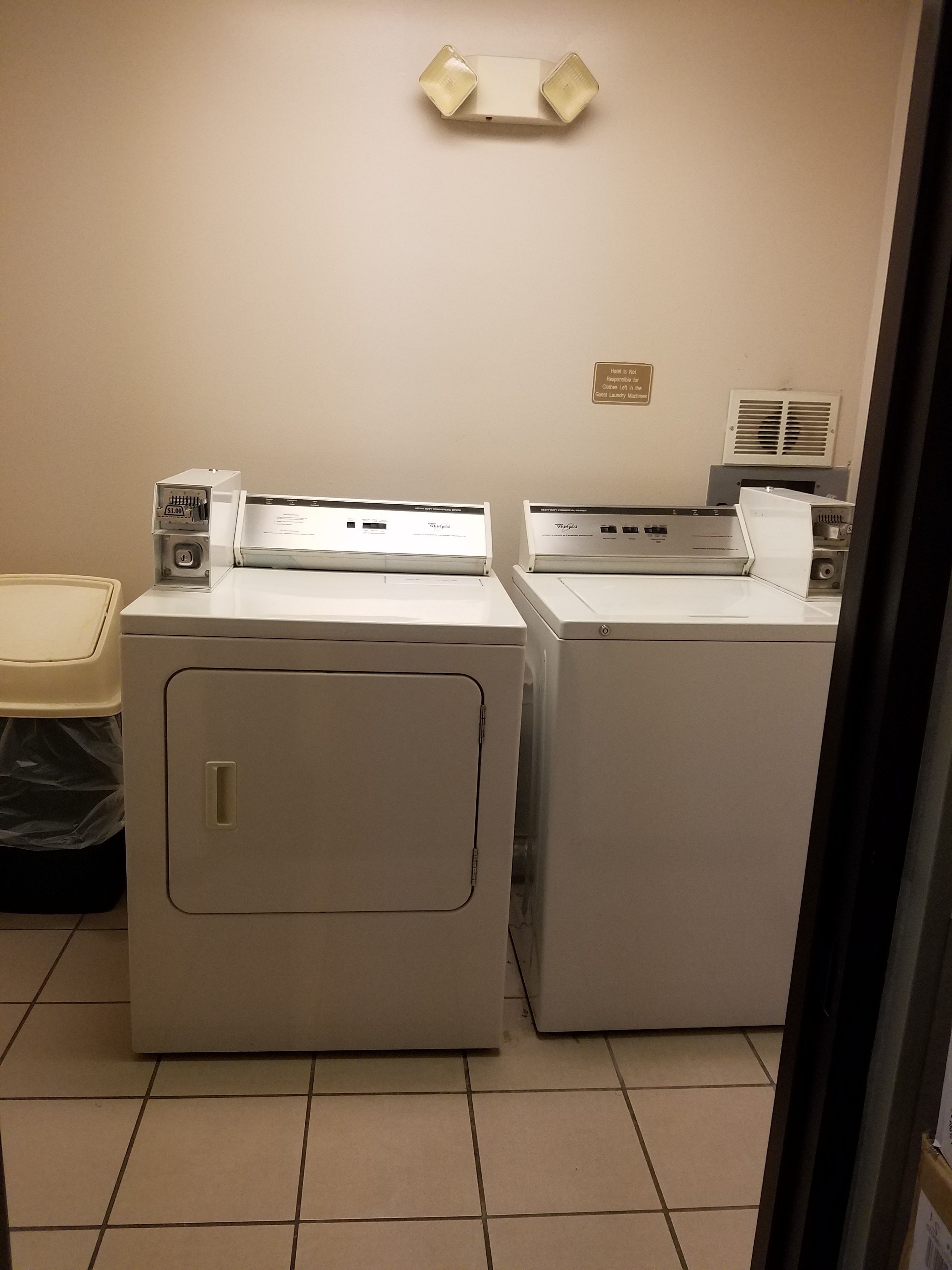 A laundry room with two front-loading washing machines and a dryer, a trash bin, and a wall-mounted light fixture.