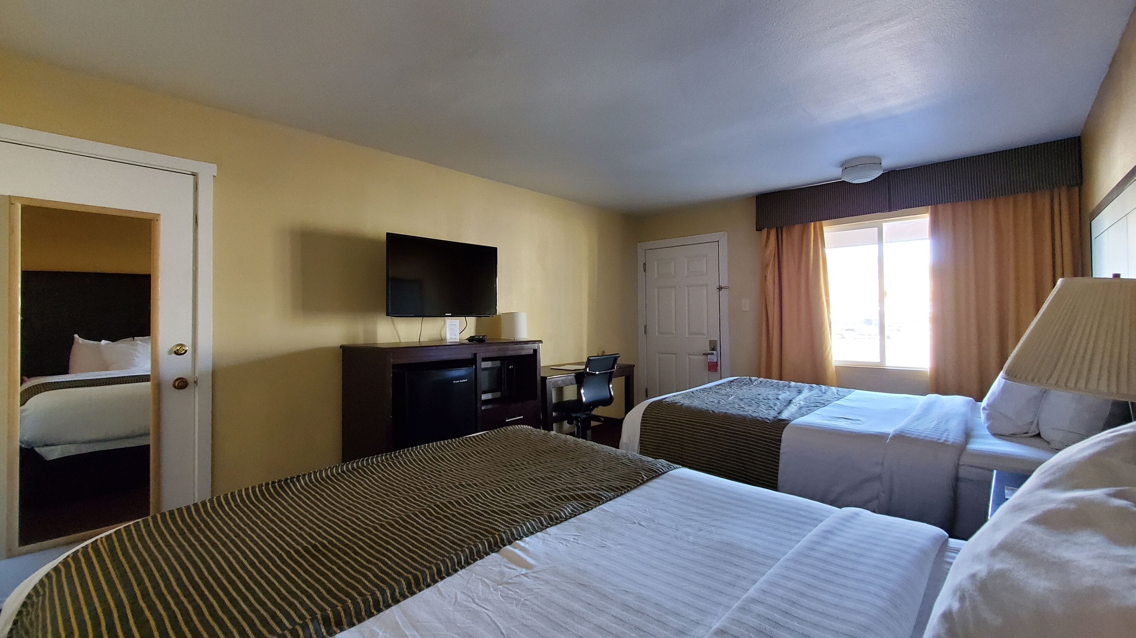 Habitación de hotel con dos camas, un escritorio con televisor, una puerta y una ventana con cortinas.