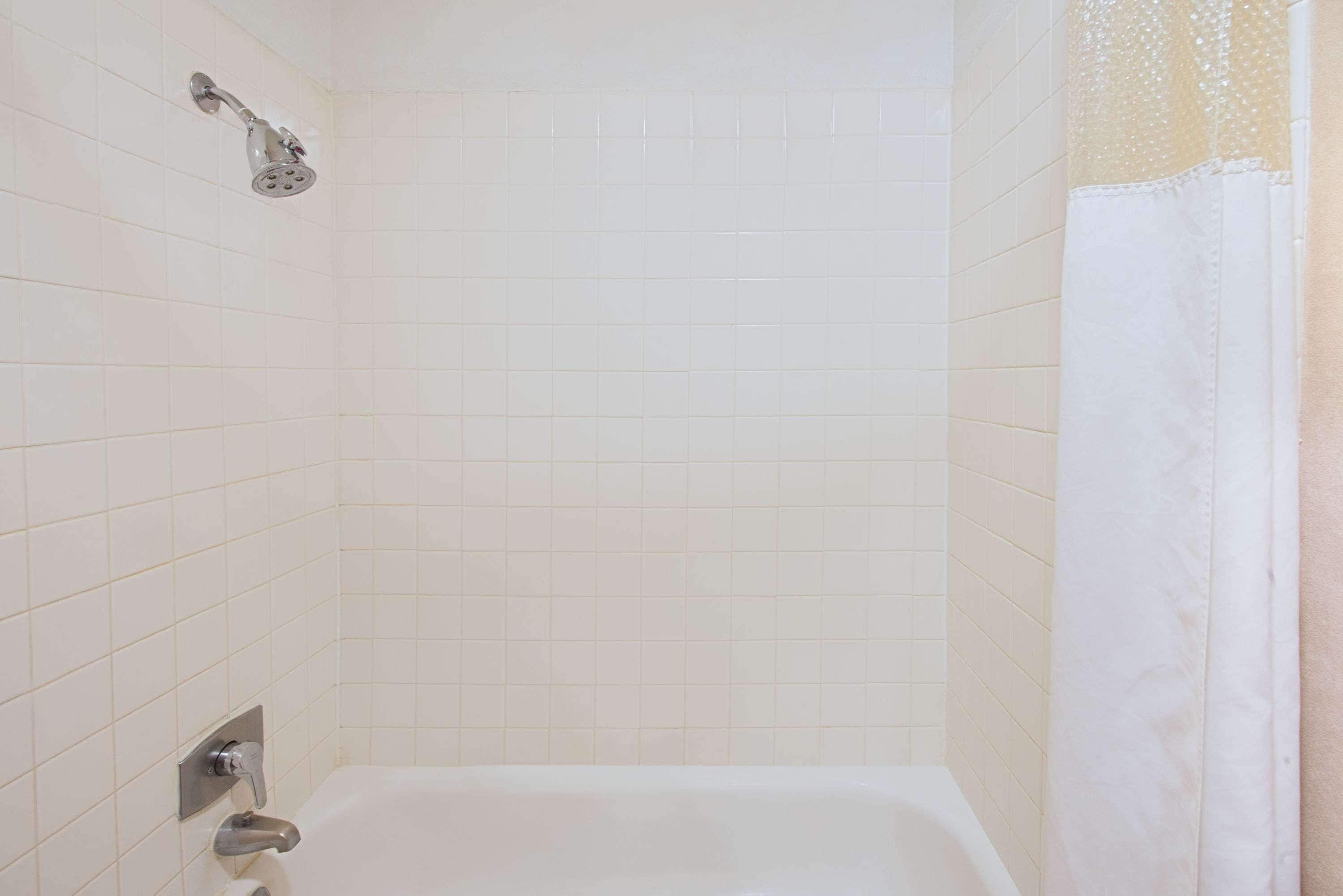 Un baño con paredes de azulejos blancos, un cabezal de ducha, una bañera y una cortina.