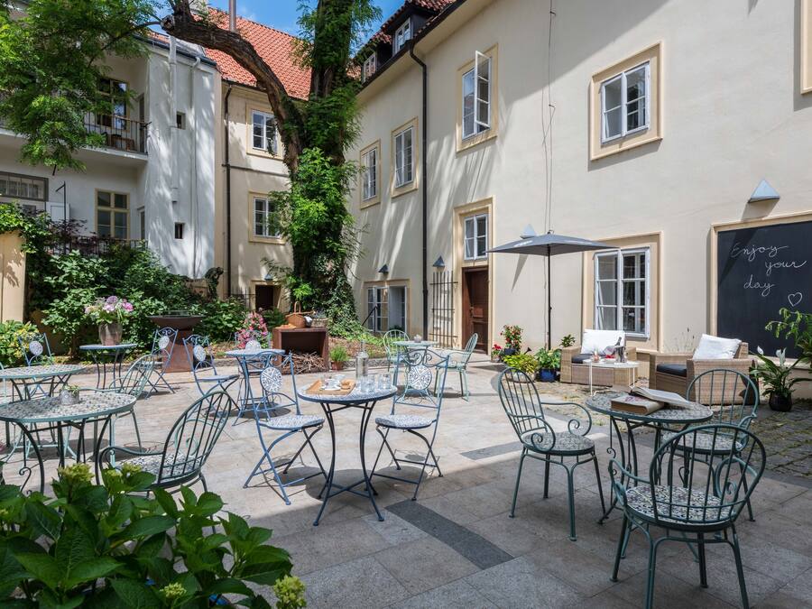 Ein Außenbereich mit Terrasse, mit mehreren Tischen und Stühlen, umgeben von Gebäuden mit Balkonen und Blumentöpfen.