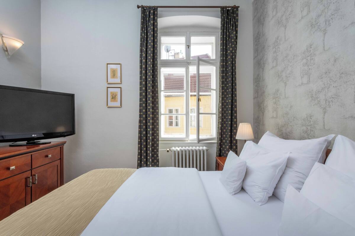A hotel room with a bed, a TV, a window with curtains, a radiator, and a lamp.