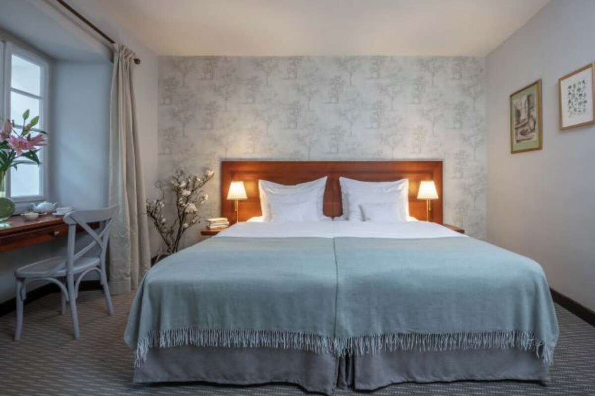 Una cama doble con cabecera de madera, dos lámparas de noche, un escritorio con silla y una ventana con cortinas.