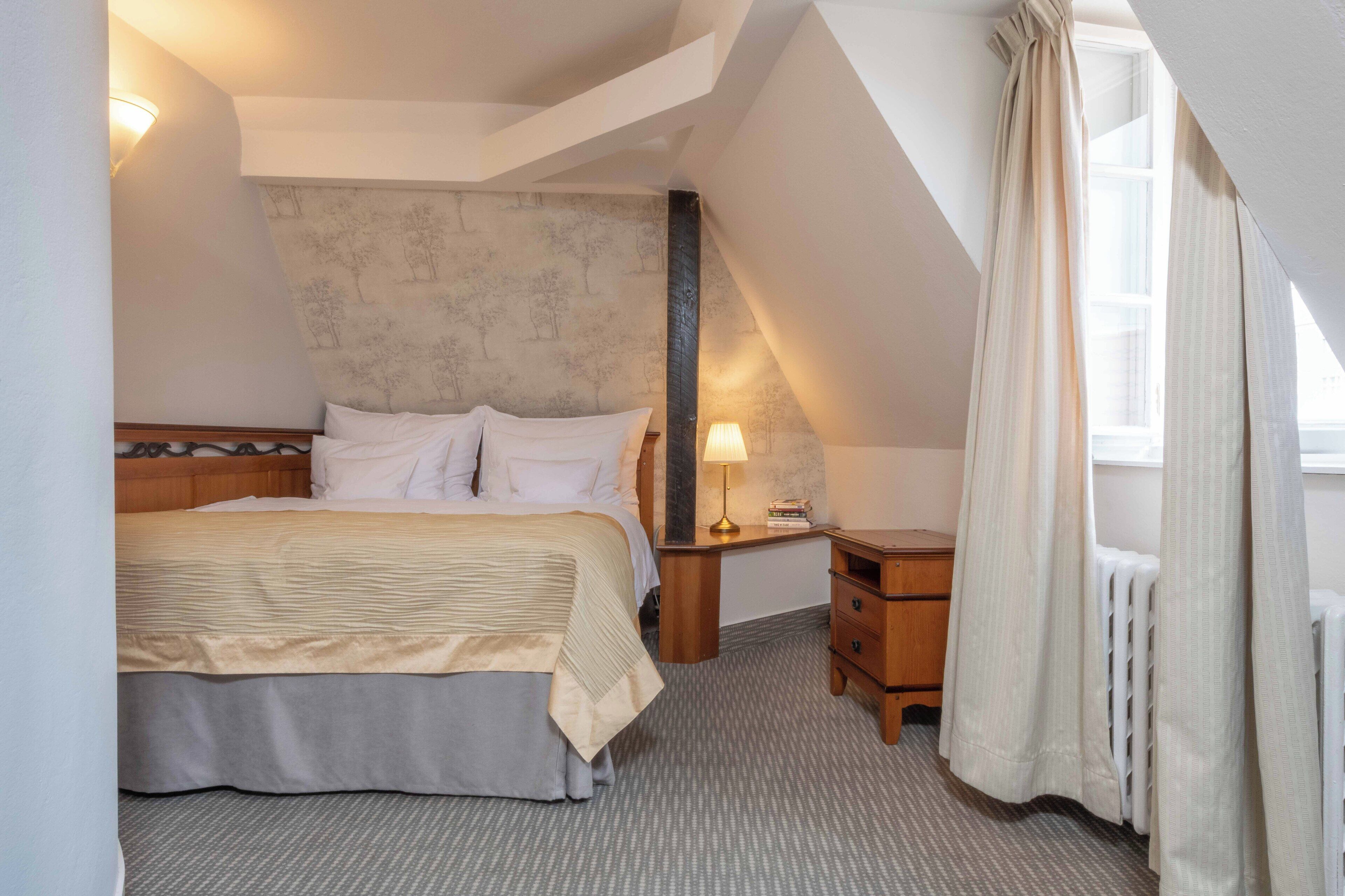 A bedroom with a bed, bedside table, lamp, and a view of the outside.